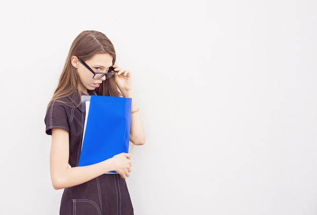 Девушка-подросток в очках с длинными волосами стоит у белой стены и держит синюю тетрадь