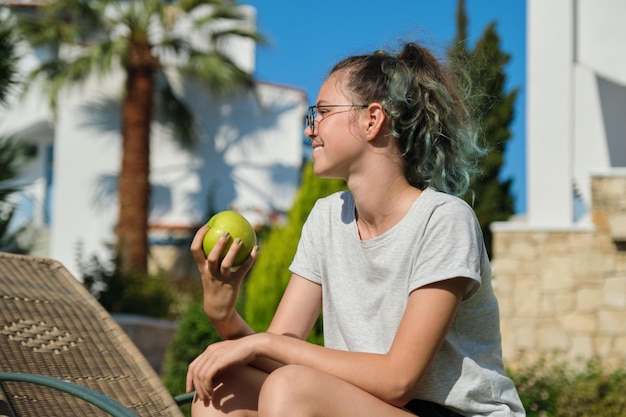정원에 있는 일광욕용 긴 의자에 앉아 녹색 사과를 들고 있는 십대 소녀