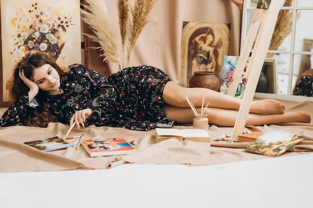 Girl in a thin black dress with a floral print lies between\
paints and paintings