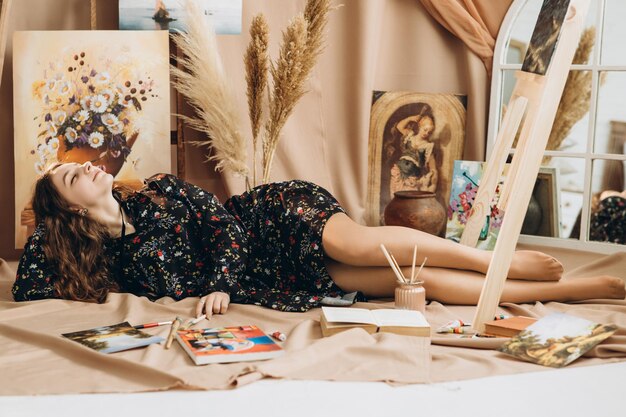 girl in a thin black dress with a floral print lies between paints and paintings