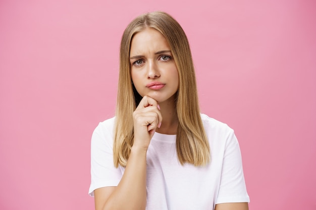 Girl thinking and holding hand on chin over pink background in white casual t-shirt.