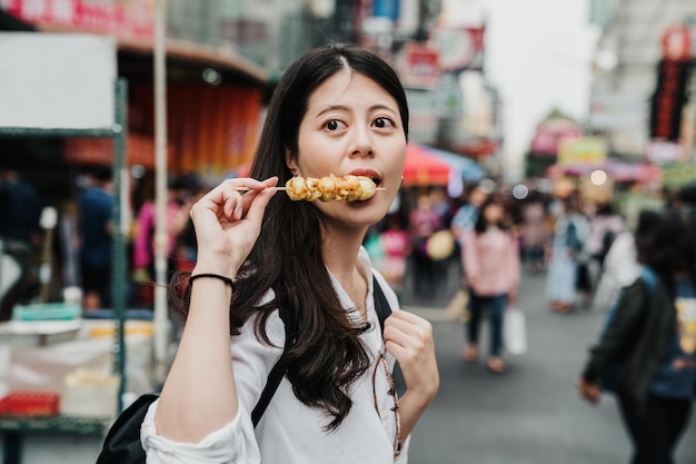 街の通りの屋外でおやつの時間に串を食べて空腹の女の子の観光客。卵の棒を持って脇を見ているアジアの日本人女性は、おいしい地元の食べ物を楽しんでいます。かわいい女の子がおいしい料理で口を開ける