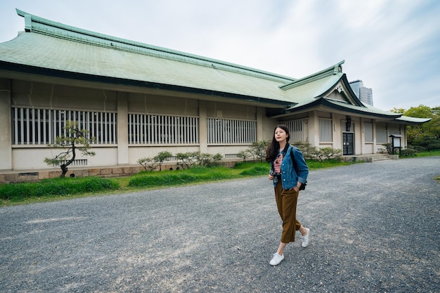 青い空を背景に大阪城を屋外でリラックスした女の子の観光客。日本での訪問ツアー中にカメラを保持しているカジュアルなスーツを着た若い女性。歩いているアジア人女性の全身写真。