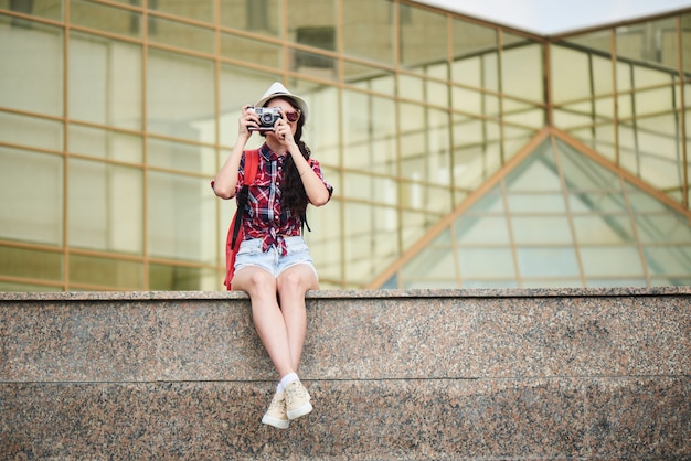 ガラスのファサードを持つ建物に対して大理石の表面に座って、都市の風景の写真を撮る女の子の観光客