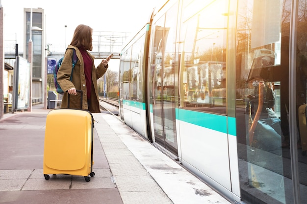 Girl tourist with a backpack and a big yellow suitcase stands on the platform and waits for the train