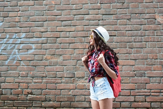 The girl tourist with hat and glasses stands on a background of wall of red brick