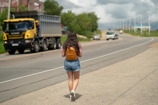 道路でヒッチハイクするバックパックを持つ女の子の観光客。ヒッチハイク観光のコンセプト。