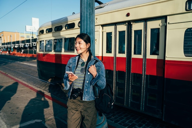 girl travel photographer get off from the light rail smiling walking back to hotel with sunset. busy urban city san francisco lifestyle concept. young asian woman holding cellphone chatting online.