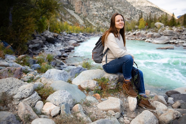 Girl traveler tourist with backpack and camera next to turquoise mountain river