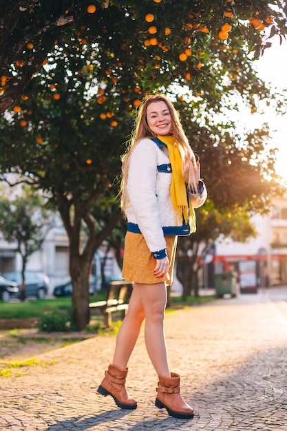 Girl in trendy clothes with long hair looks at camera and smile. city street and orange trees.