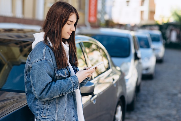 車にもたれて、電話で何かを見て、都市の背景に女の子