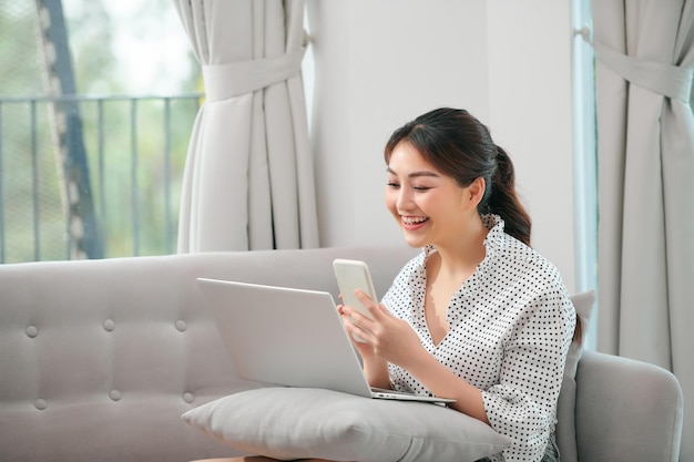 Girl using laptop and smartphone Young woman in casual clothes with her legs crossed sitting on sofa at cozy home interior Technology and communication concept