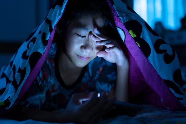 Girl using mobile phone on dark bed in the bedroom