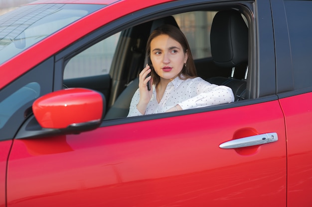 La ragazza che per mezzo dello smart phone e l'attesa dell'alimentazione elettrica si collegano ai veicoli elettrici per caricare la batteria in automobile. la ragazza positiva che parla sul telefono si siede in automobile elettrica e nella carica.