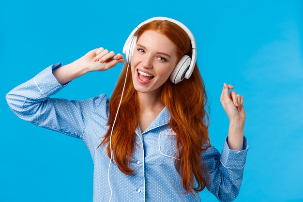 Girl waking up starting day with music. Joyful carefree redhead teenager dancing in nightwear, raising hands up wearing large white headphones singing along favorite song, playlist, blue background