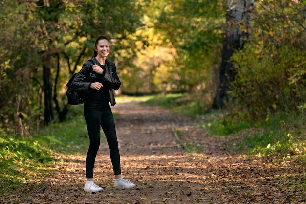 Girl walks along the alley of the autumn park. Happy girl in black clothes in the forest. Alley, promenade. Sunny autumn day.