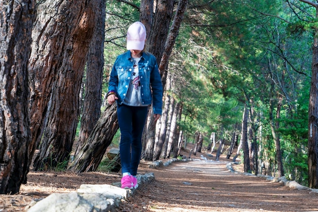 A girl walks along the curb with her head down in a pine forest on a sunny path.