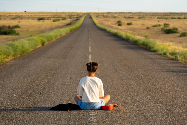 A girl walks along an empty road in the steppe she has a\
ukulele and a backpack open road