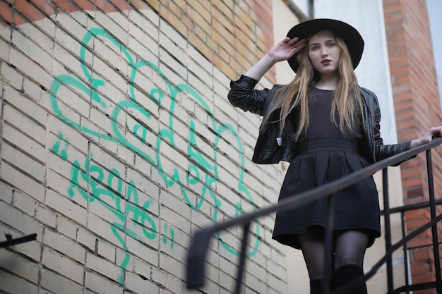 A girl walks down the city street in a leather waistcoat with a phone. Young beautiful girl in a hat and with a dark make-up outside. Girl in the Gothic style on the street.