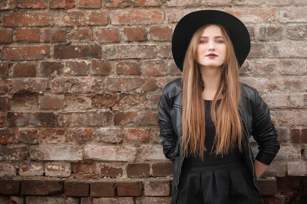 A girl walks down the city street in a leather waistcoat with a phone Young beautiful girl in a hat and with a dark makeup outside Girl in the Gothic style on the streetxA