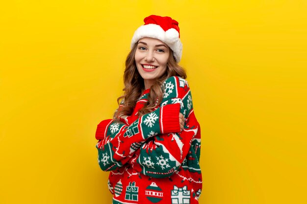 Girl in warm christmas sweater and santa claus hat hugs herself on yellow isolated background