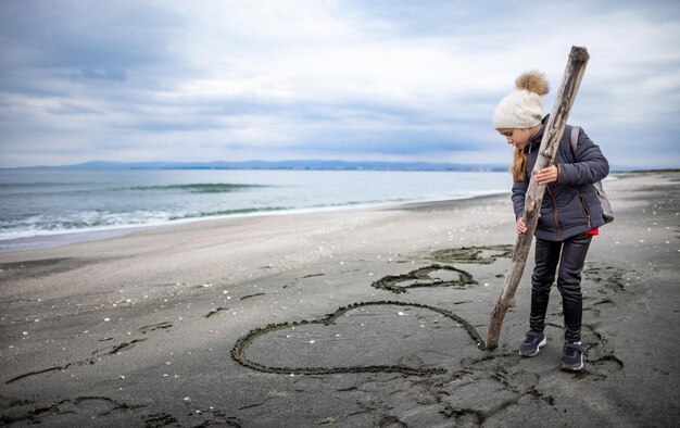 A girl in warm clothes and a hat with a backpack walks along the beach and draws drawings and symbols on the sand