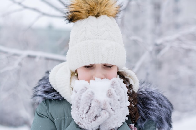 暖かい服を着た女の子が冬の公園で手袋をはめて雪を手に持っている冬のライフスタイルの肖像画
