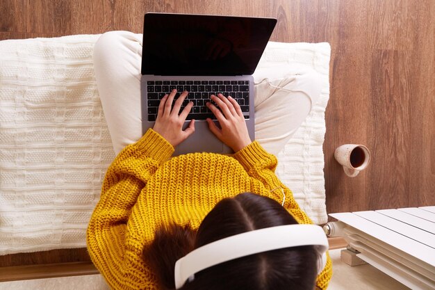 A girl in warm clothes is sitting on the floor in the house by the battery and working on a laptop Distance education The concept of a cold winter problems with central heating Top view