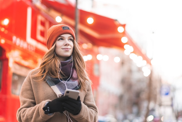 girl in warm clothes stands in the street with a smartphone in her hands, listens to music in the headphones