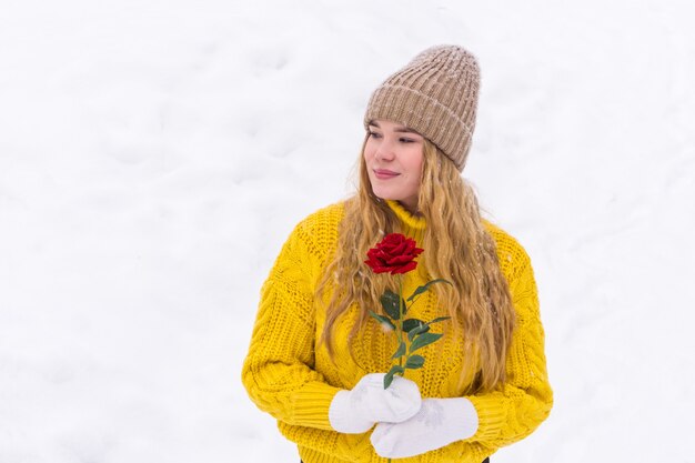 Girl in a warm sweater and knitted hat holds a rose in her hands against a snowy background