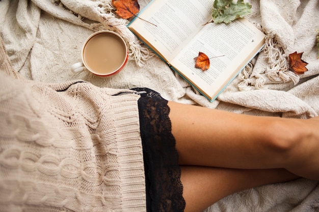 A girl in a warm sweater sits on a beige woolen blanket and holds a mug of hot coffee in her hands. 