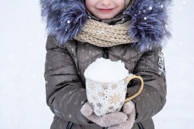 暖かい冬のアウトドア服を着た女の子が手に雪の入ったマグカップを持っている寒い天候の季節性クリスマスと新年