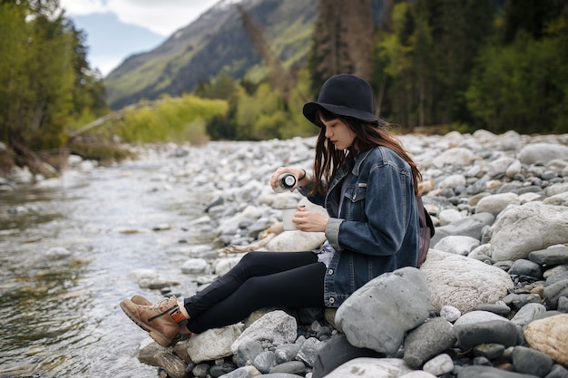Девушка в рюкзаке пьет чай из термоса и смотрит на озеро