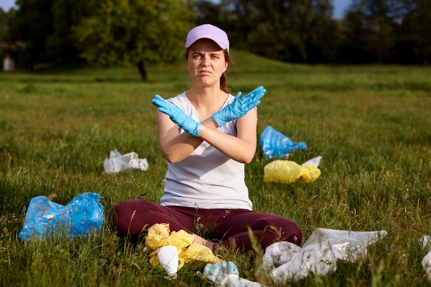 Girl wearing baseball cap and casual shirt, sitting on green grass and crossing hands, calls for do not pollute planet, surrounded with rubbish, being upset, solving ecological problems.