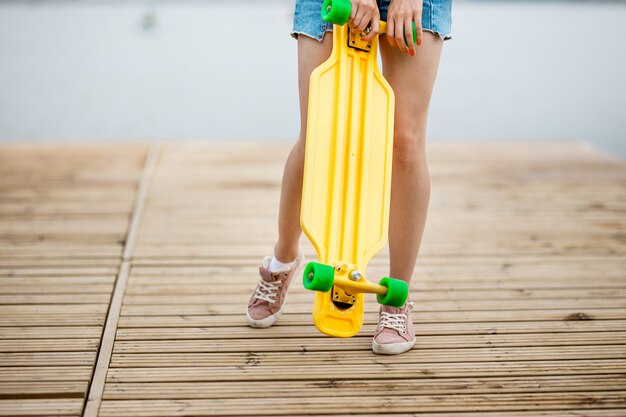 a girl wearing denim shorts is standing on the pier and holding a yellow longboard in front of her.