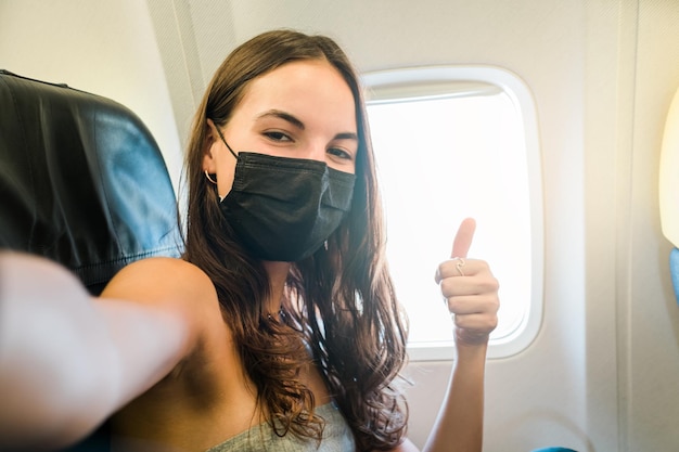 Девушка в маске делает селфи со смартфоном, сидя в самолете
