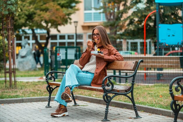 Una ragazza che indossa occhiali e abiti alla moda si siede su una panchina nel parco e mangia deliziosi dolci messa a fuoco selettiva