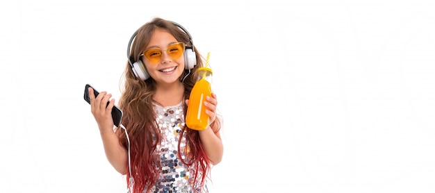 Girl wearing glittery dress and yellow sunglasses, with big white earphones, listening to music and holding black smartphone and bottle with orange juice isolated