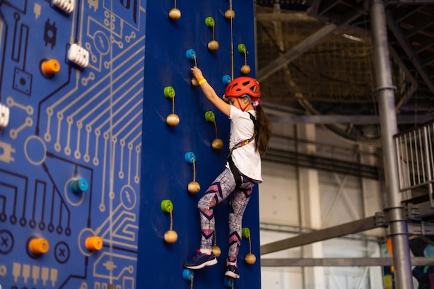 Girl wearing in harness and safety equipment climbing on practical wall indoor. Girl inserting the rope in a quickdraw.