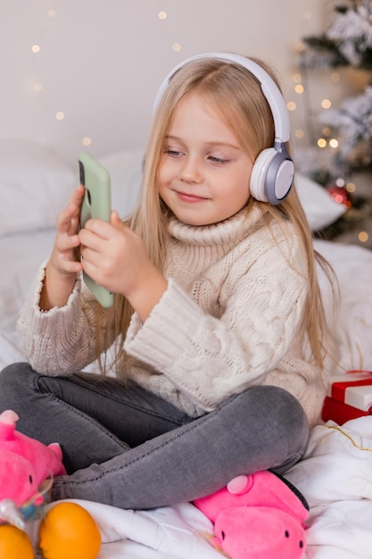 A girl wearing headphones sits on a bed with a christmas tree in the background.