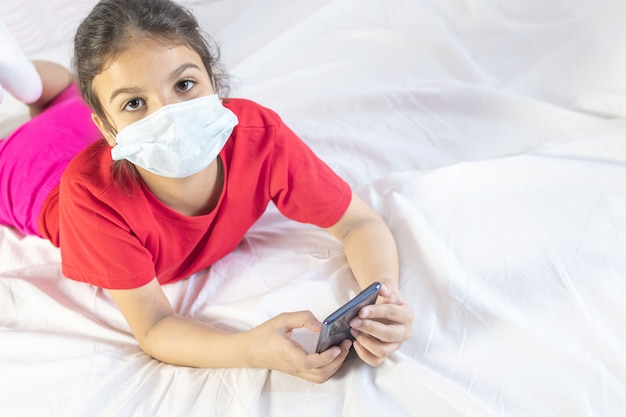 Girl wearing mask, using mobile for school or games. Laying on bed with mobile.