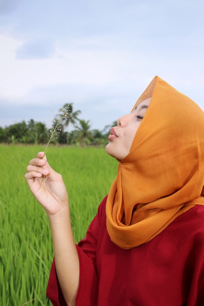 Foto ragazza che indossa un hijab arancione e soffia un fiore in uno sfondo di campo