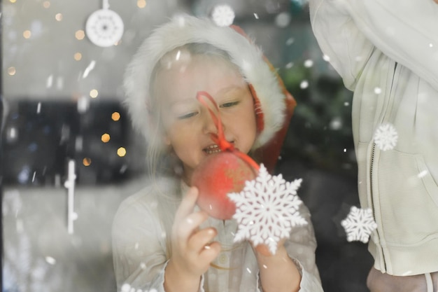 A girl wearing a santa claus hat looks at snowflakes on the window