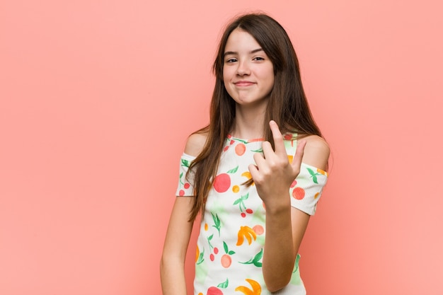 Girl wearing a summer clothes against a red wall pointing with finger at you as if inviting come closer.