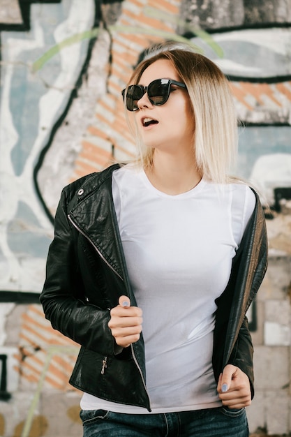 Girl wearing t-shirt, glasses and leather jacket posing against street