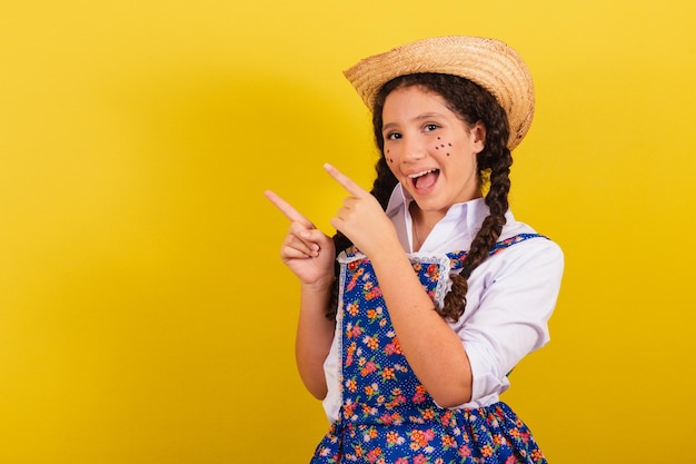 Girl wearing typical clothes for Festa Junina pointing to the side with fingers space for ad and text For the Arraia party