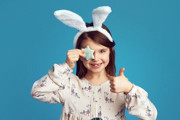 Girl wears bunny ears covers eye with star shape cookie and shows thumb up