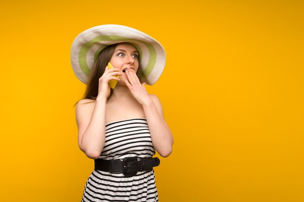 Girl wears straw hat and short white strips dress with open shoulders talking on a smartphone