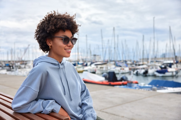 La ragazza indossa occhiali da sole e felpa con cappuccio si diverte a navigare in barca a vela e in barca a vela durante le passeggiate nel porto di mare durante il giorno ha viaggi di lusso