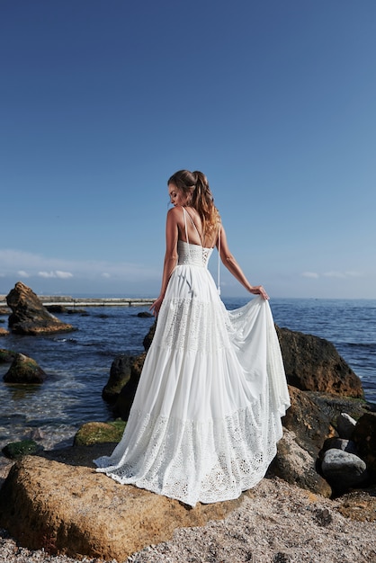 Girl in wedding luxury dress posing on sea shore. Bride on a rocks.